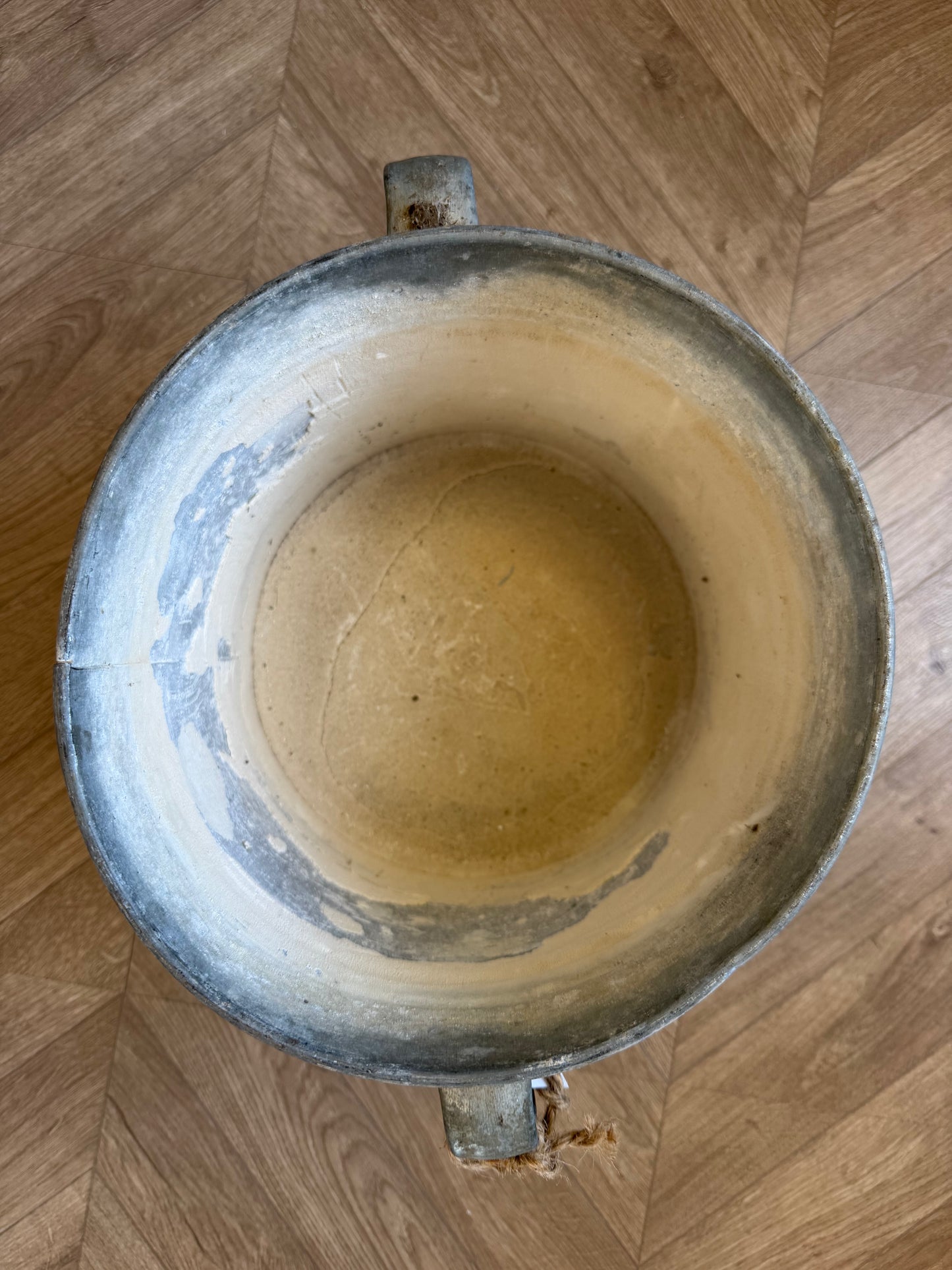 A top-down view of a large, vintage-style metal bucket with a distressed silver-gray exterior and a worn, yellowish-beige interior, set on a light brown wooden parquet floor.